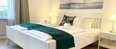 Una cama bien hecha con un plaid verde, ropa de cama blanca y un cabecero con un cojín floral. Hay dos mesitas de noche con lámparas y una ventana con cortinas.