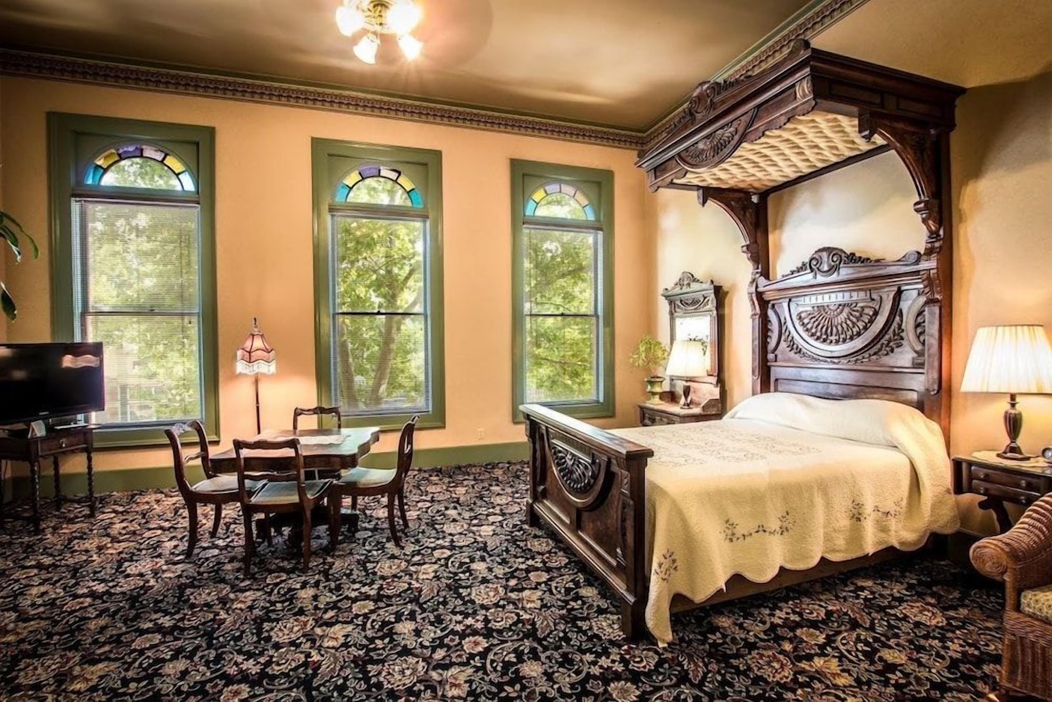 A bedroom with a four-poster bed, a television, a desk with a chair, and a window with a view of trees.