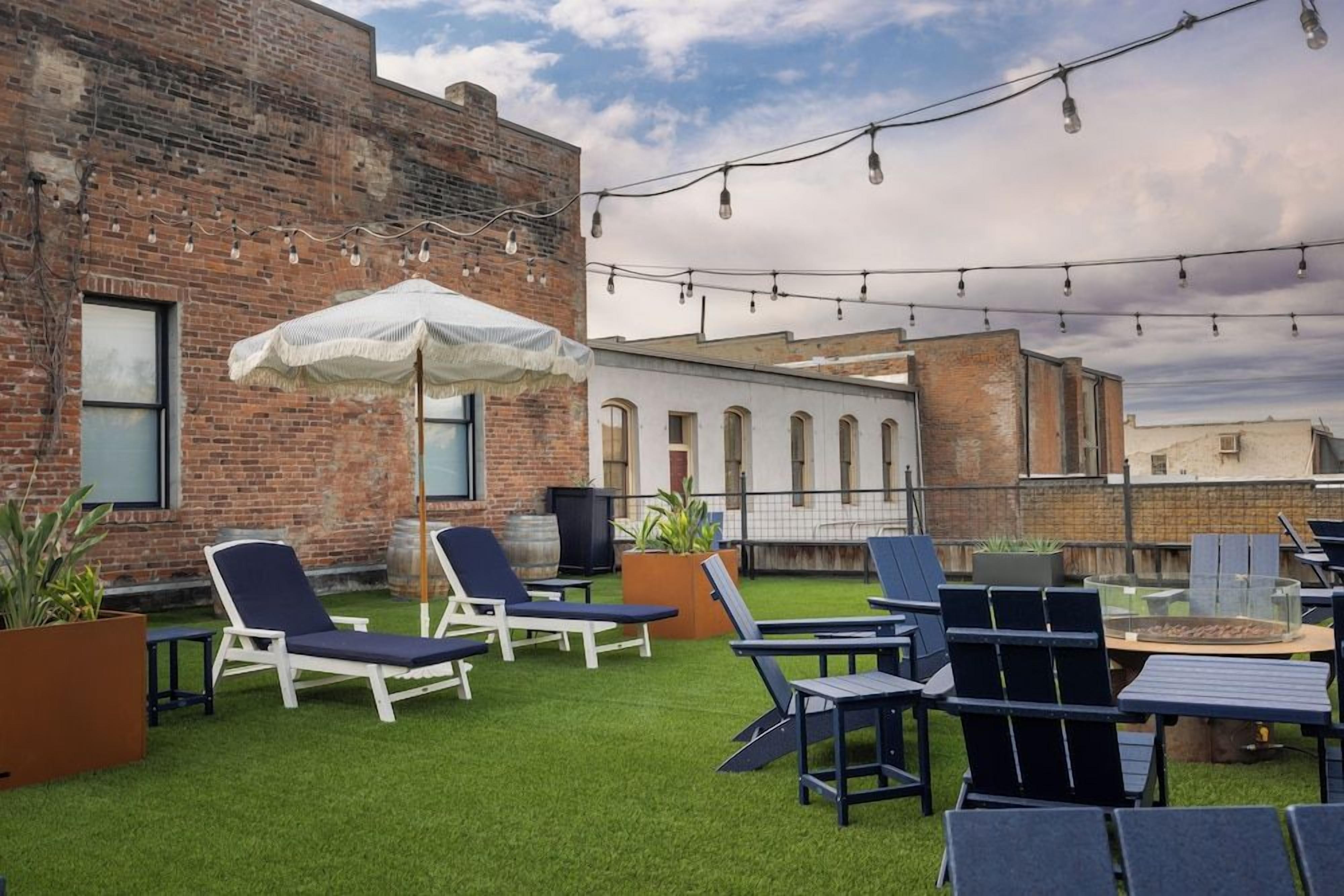 An outdoor seating area with lounge chairs and tables, a white umbrella, and string lights against a brick building backdrop.