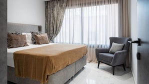 A modern bedroom with a large bed, a grey armchair, and a view of the outdoors through sheer curtains. - Casino & Mall Jacuzzi Penthouse (Lemesos)