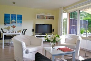 Un salon comprenant un téléviseur, deux fauteuils blancs, une table basse ornée d’un vase de fleurs et un coin repas avec une table et des chaises.