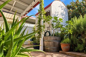 A covered outdoor area with a white building, potted plants, and a wooden barrel. - Tucked away in a quiet pocket near Fremantle, this character-filled private garden cottage comfortably sleeps 2. Enjoy morning coffee on the sunny front verandah, relax among lavender and vines in the leafy fenced garden, and stroll into Freo’s cafés (Beaconsfield)