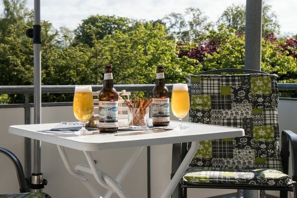 A table with two bottles and two glasses of beer, a chair with a patterned cushion, and a railing with greenery in the background.
