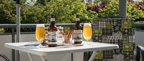 Une table avec deux bouteilles et deux verres de bière, une chaise avec un coussin à motifs et, en arrière-plan, un garde-corps orné de plantes.