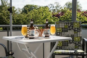A table with two bottles and two glasses of beer, a chair with a patterned cushion, and a railing with greenery in the background. - Studio 11 on the 1st floor of Haus Stranddistel (Zingst)