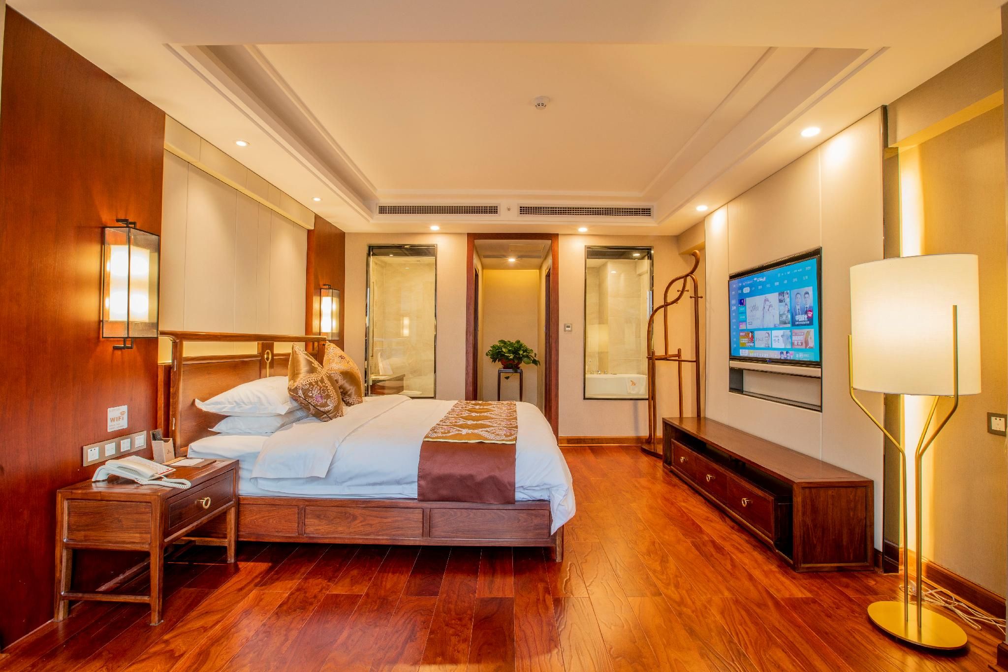 Habitación de hotel con piso de madera, cama con ropa blanca, mesita de noche con teléfono, televisor en la pared y lámpara.