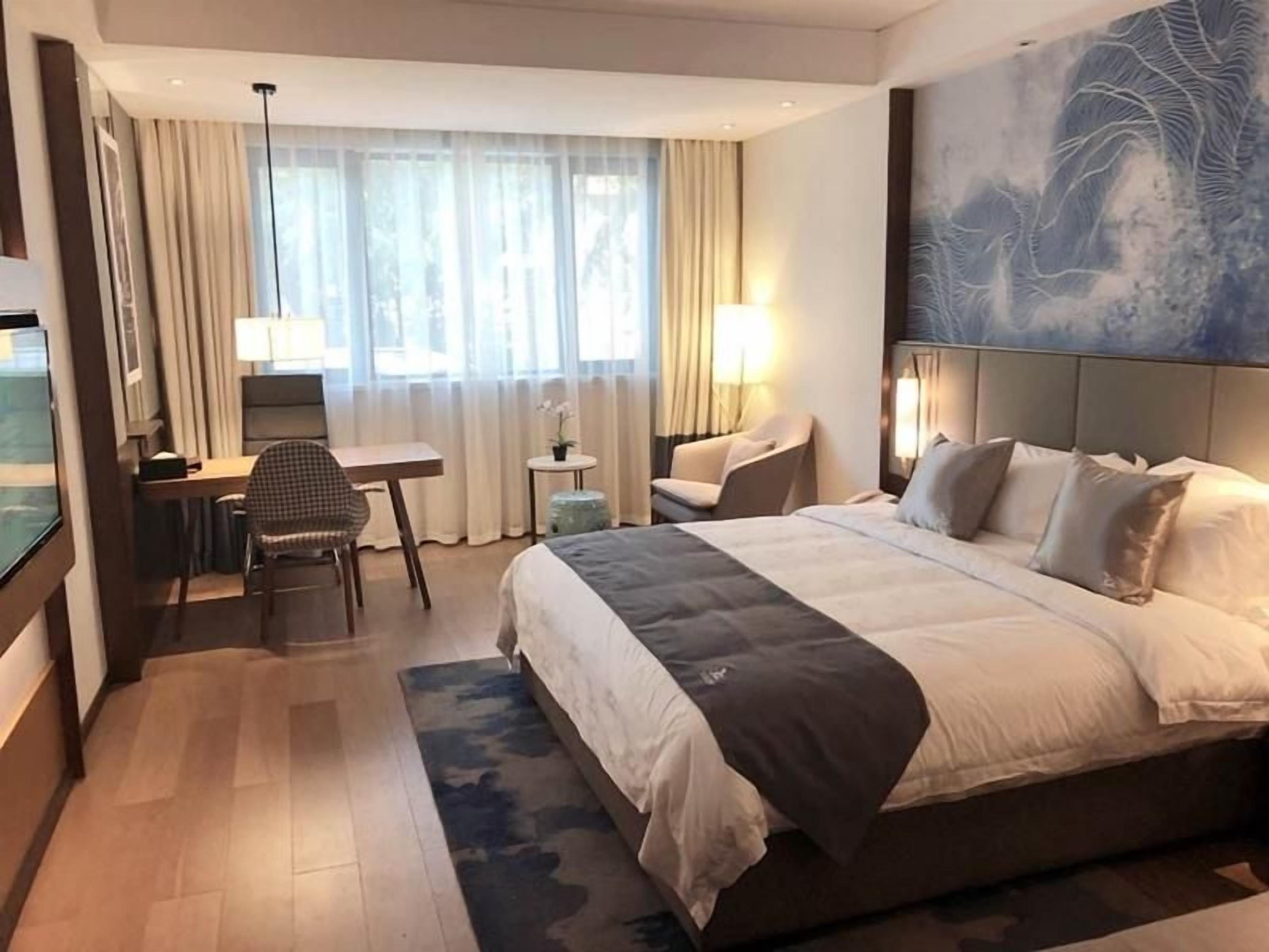 Una habitación de hotel con cama, escritorio, silla y un ventanal con cortinas.