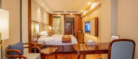 A hotel room with a bed, a desk, a chair, a small table, and a television.