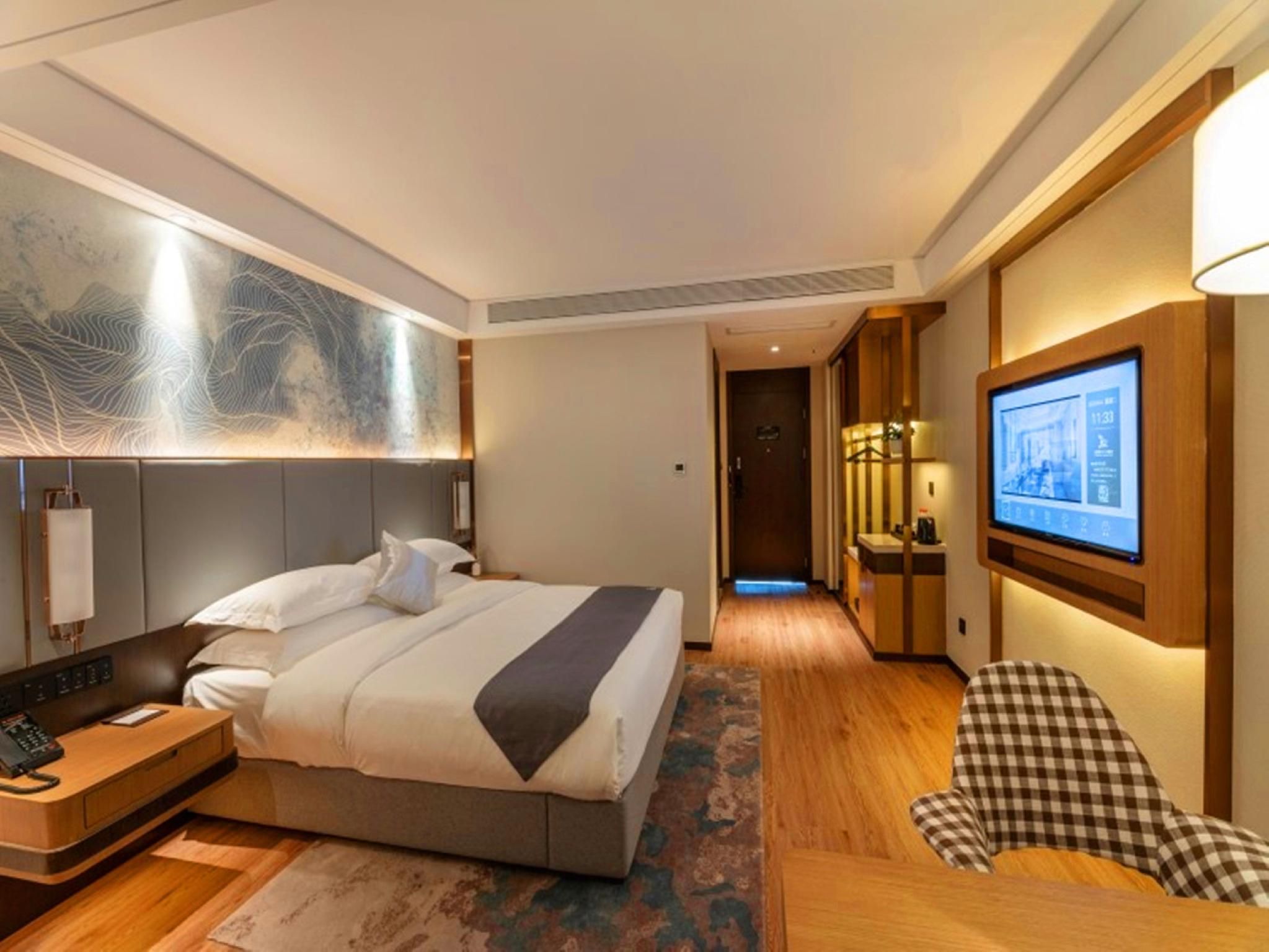 A hotel room with a bed, bedside table, lamp, artwork, television, and a checkered chair.