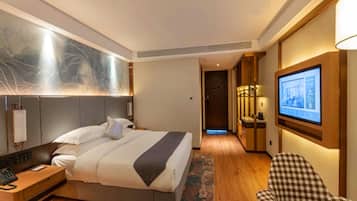 A hotel room with a bed, bedside table, lamp, artwork, television, and a checkered chair.