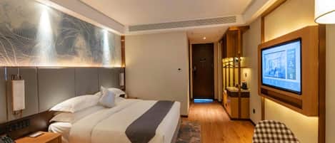 A hotel room with a bed, bedside table, lamp, artwork, television, and a checkered chair.
