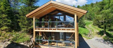 A two-story wooden cabin with a glass balcony, surrounded by forest.