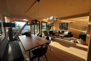 A spacious living area with a wooden dining table, black chairs, a sofa, and a coffee table. There are large windows providing natural light and a view of the outdoors.