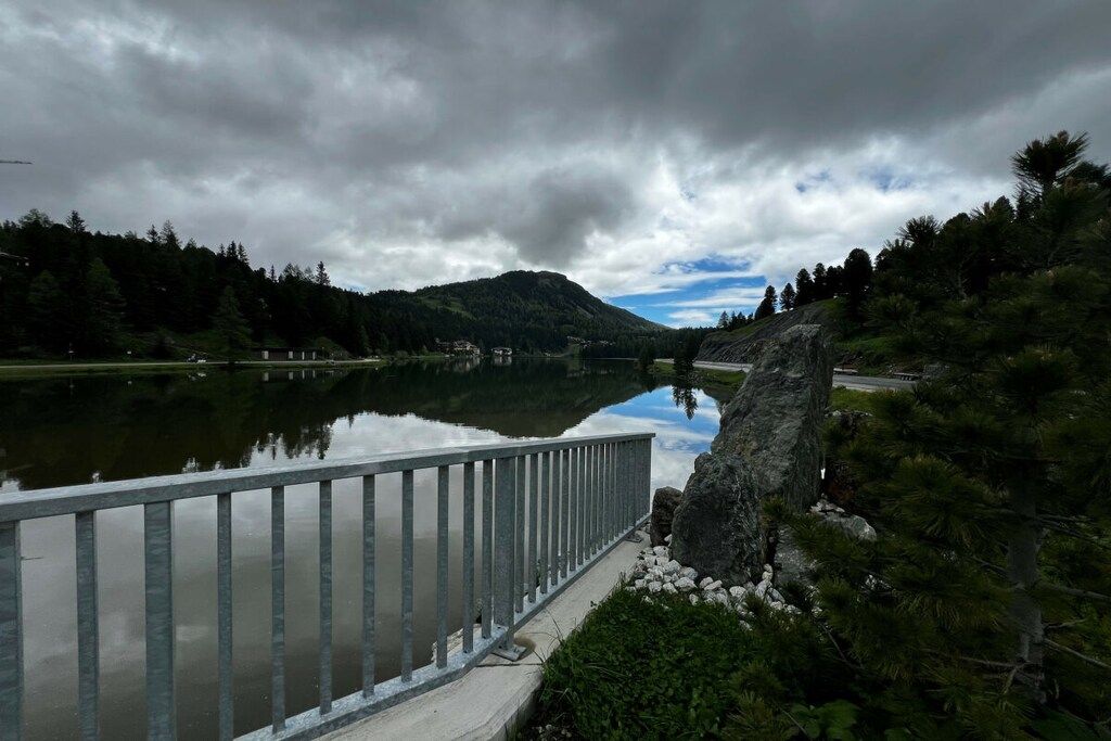 Um lago calmo com guarda-corpo metálico, rodeado por uma floresta densa e uma montanha ao fundo.