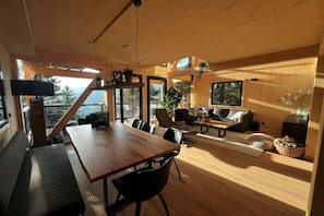 A spacious living area with a wooden dining table, black chairs, a sofa, and a coffee table. There are large windows providing natural light and a view of the outdoors.