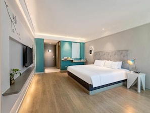 A modern hotel room with a large bed, a bedside table, a TV mounted on the wall, and a bathroom visible through an open door.