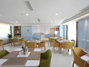 A modern dining area with multiple tables and chairs, a kitchen in the background, and a bookshelf.