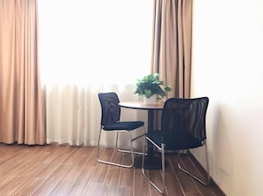 Una stanza con pavimento in legno, due sedie nere, un piccolo tavolo rotondo, una pianta in vaso e tende color beige.