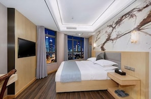 A hotel room with a bed, a TV, a nightstand, a window with a city view, and a wall with a tree branch design. - Junting (Chongqing)