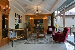 A spacious living area with a fireplace, a chandelier, and a large window providing a view of the outdoors. - Micheline Hotel Tricot (Gramado)