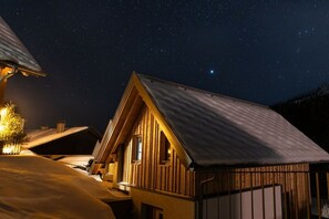 Eine hölzerne Hütte mit geneigtem Dach, umgeben von Schnee, unter einem sternenübersäten Nachthimmel.