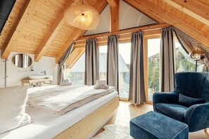Ein Schlafzimmer mit Holzboden, einem Bett, einem Sessel, einem Nachttisch und einem Fenster mit Vorhängen.