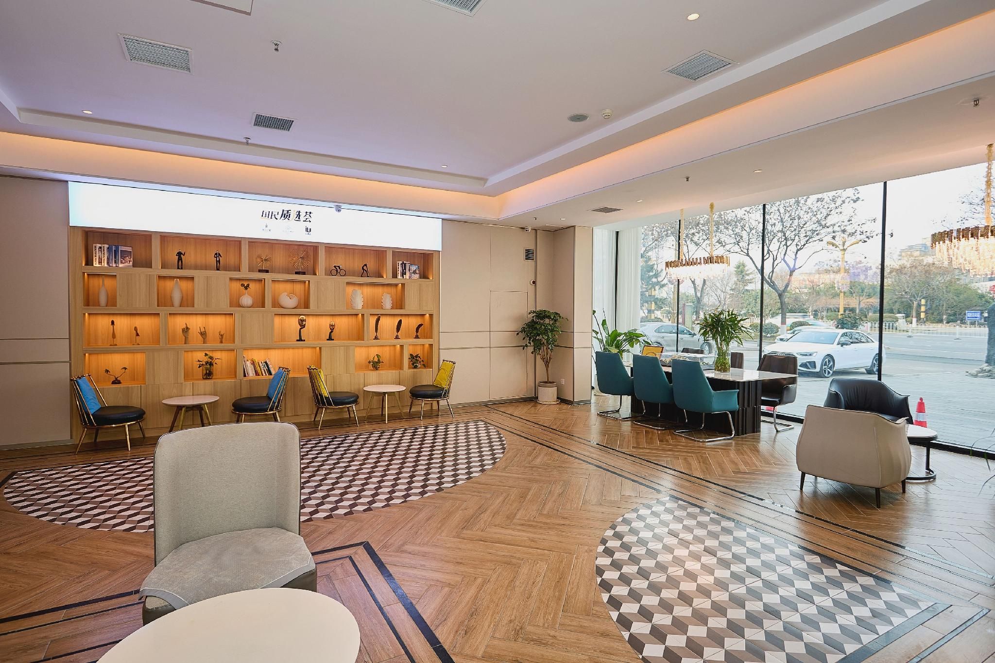 A modern hotel lobby with a wooden reception desk, seating area, and large windows offering a view of the street.