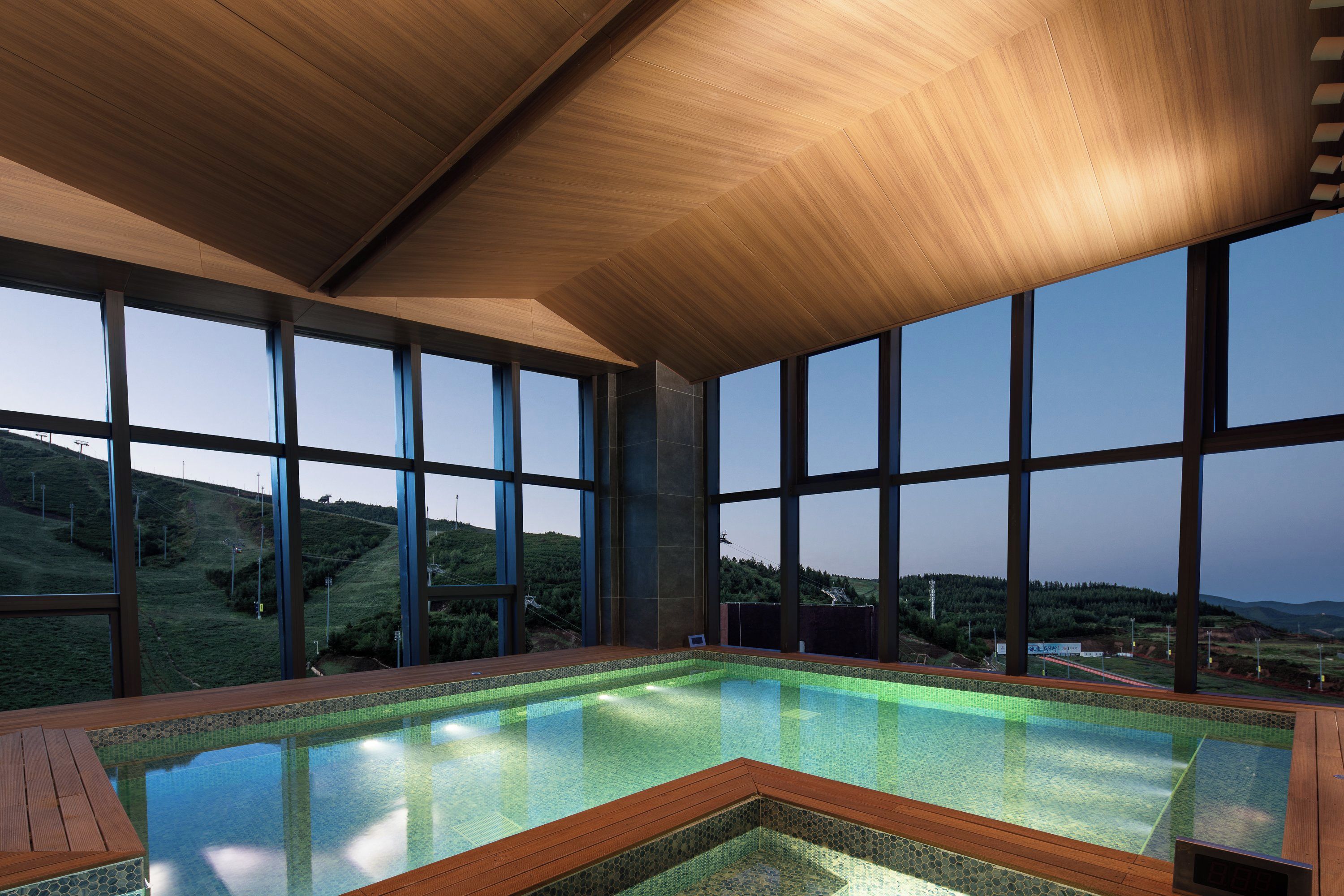 A large indoor swimming pool with a wooden deck, surrounded by large windows offering a view of a hilly landscape.