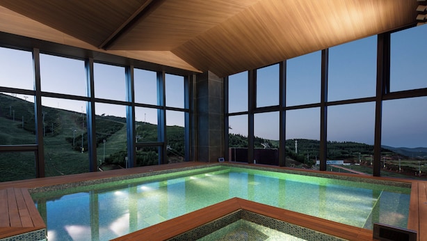 Uma grande piscina coberta com deck de madeira, rodeada por amplas janelas que oferecem vista para uma paisagem montanhosa.