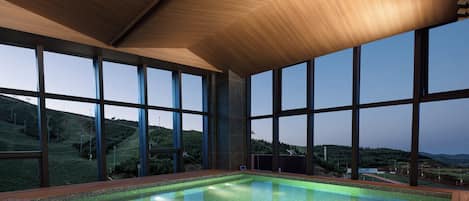 A large indoor swimming pool with a wooden deck, surrounded by large windows offering a view of a hilly landscape.