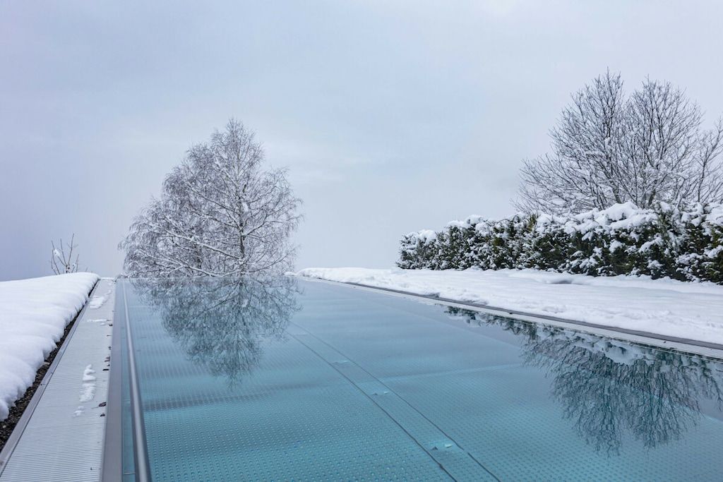 A clear swimming pool with snow-covered trees and bushes around it.