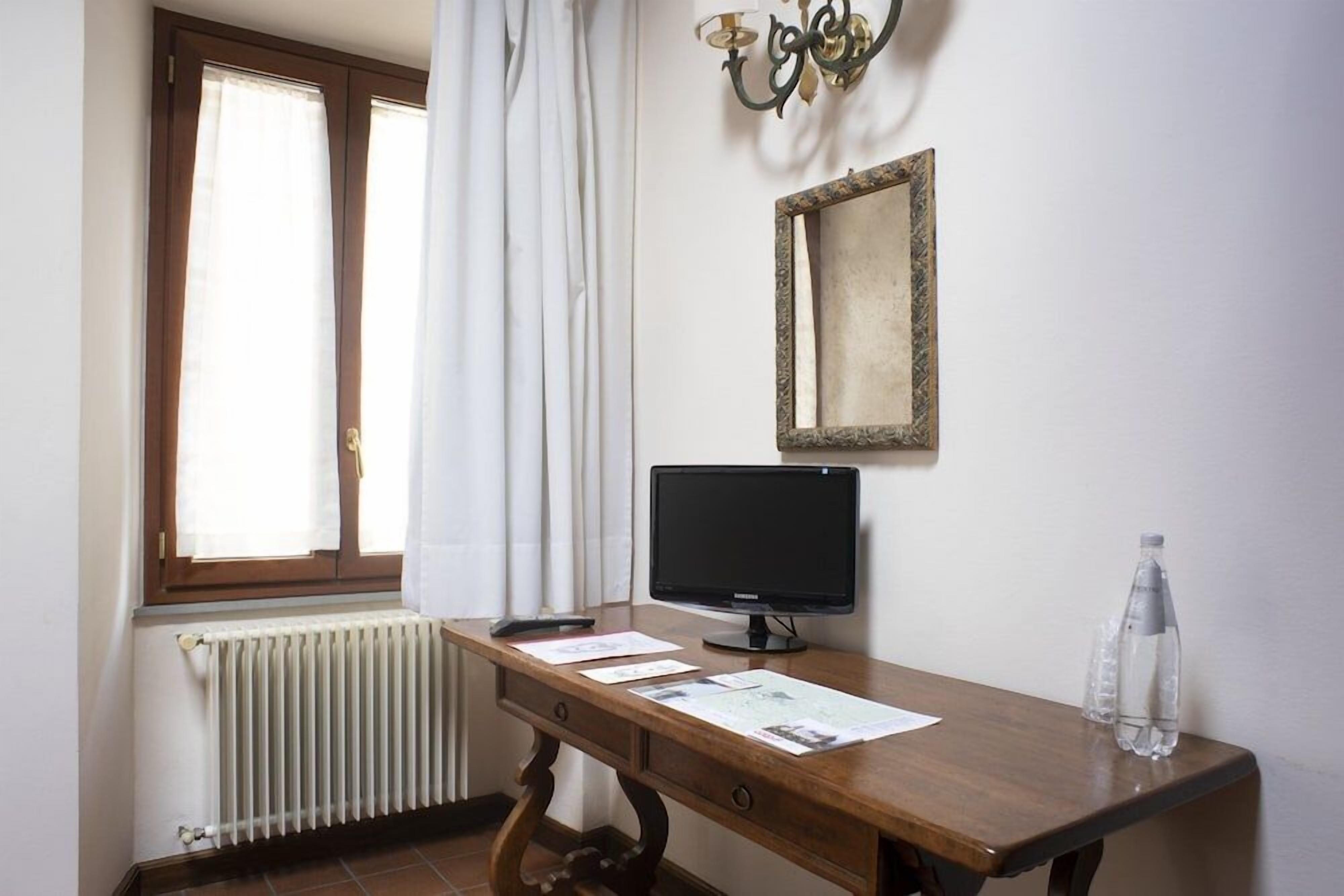 A room with a wooden desk, a black monitor, a radiator, a window with curtains, and a wall-mounted light fixture.