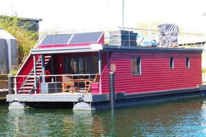A red houseboat with a deck, stairs, and a small table and chair set.