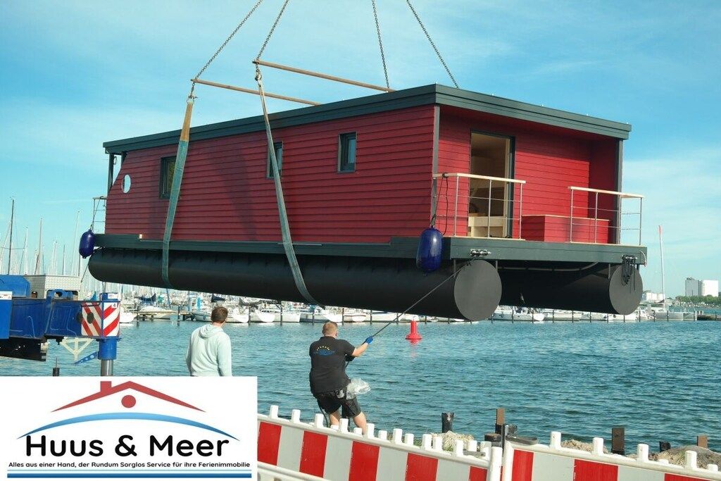 Una casa flotante roja, elevada por una grúa sobre el agua, con una persona de pie en el muelle.