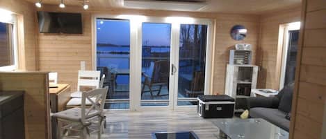 A wooden interior with a sofa, chairs, a table, a TV, and a view through glass doors to the outside.