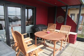 En utomhusveranda med ett runt träbord och stolar, en grill och utsikt över vattnet.