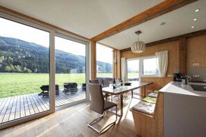 A modern kitchen with a large island, wooden beams, and a view of a green landscape through large windows. - Cabin in Murau with sauna and terrace (Murau)
