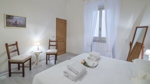 A hotel room with a bed, two chairs, a small table with a lamp, a window with curtains, and a framed picture on the wall. - Pontormo (Florence)