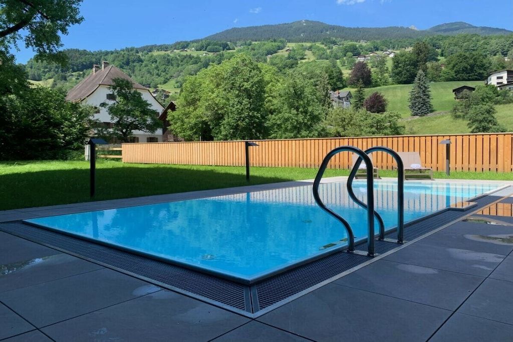 A rectangular swimming pool with a wooden fence and metal ladder, surrounded by a grassy area and a view of a hilly landscape.