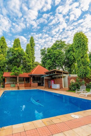 Una piscina con solárium empedrado, rodeada de árboles y un edificio con techo rojo.