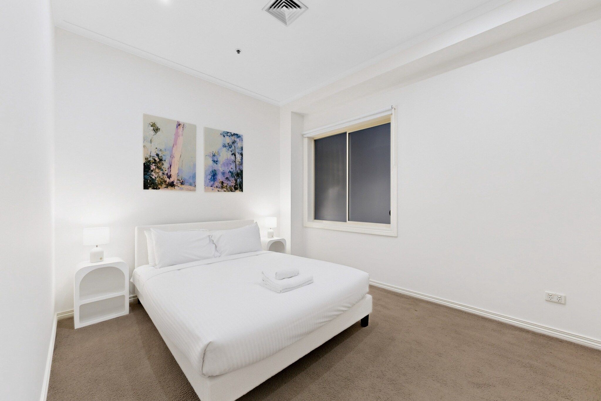 A bedroom with a bed, two bedside tables, a window, and two wall-mounted paintings.