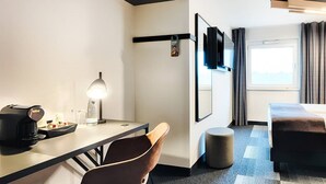 A hotel room with a bed, a desk with a coffee maker, a chair, a round ottoman, a TV mounted on the wall, and a window with curtains. - Ibis Budget Wiesbaden Nordenstadt (Wiesbaden)