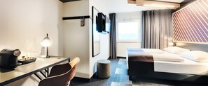 A hotel room with a bed, a desk with a coffee maker, a chair, a round ottoman, a TV mounted on the wall, and a window with curtains. - Ibis Budget Wiesbaden Nordenstadt (Wiesbaden)