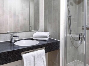A modern bathroom with a round sink, a mirror, a towel rack, and a glass-enclosed shower area. - Ibis Budget Wiesbaden Nordenstadt (Wiesbaden)