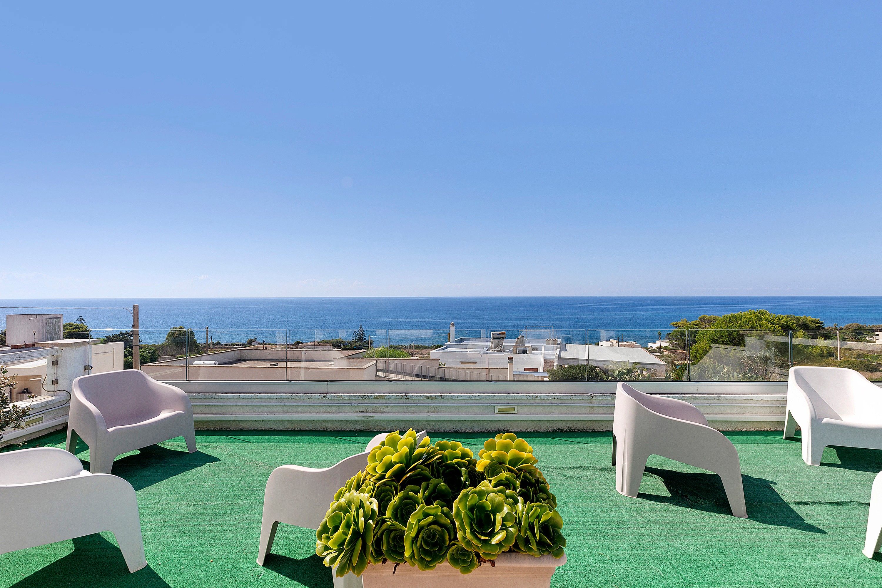 A terrace with a view of the sea, white plastic chairs, and a table with green plants.