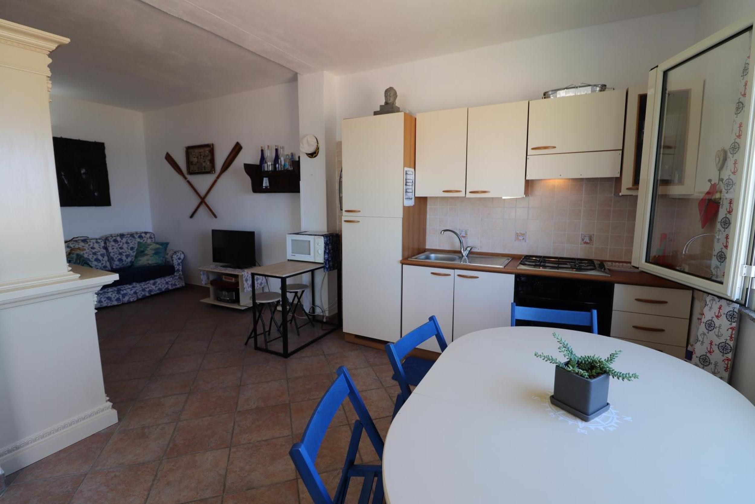 A compact kitchen with white cabinets, a dining table with blue chairs, a small TV, and a refrigerator.