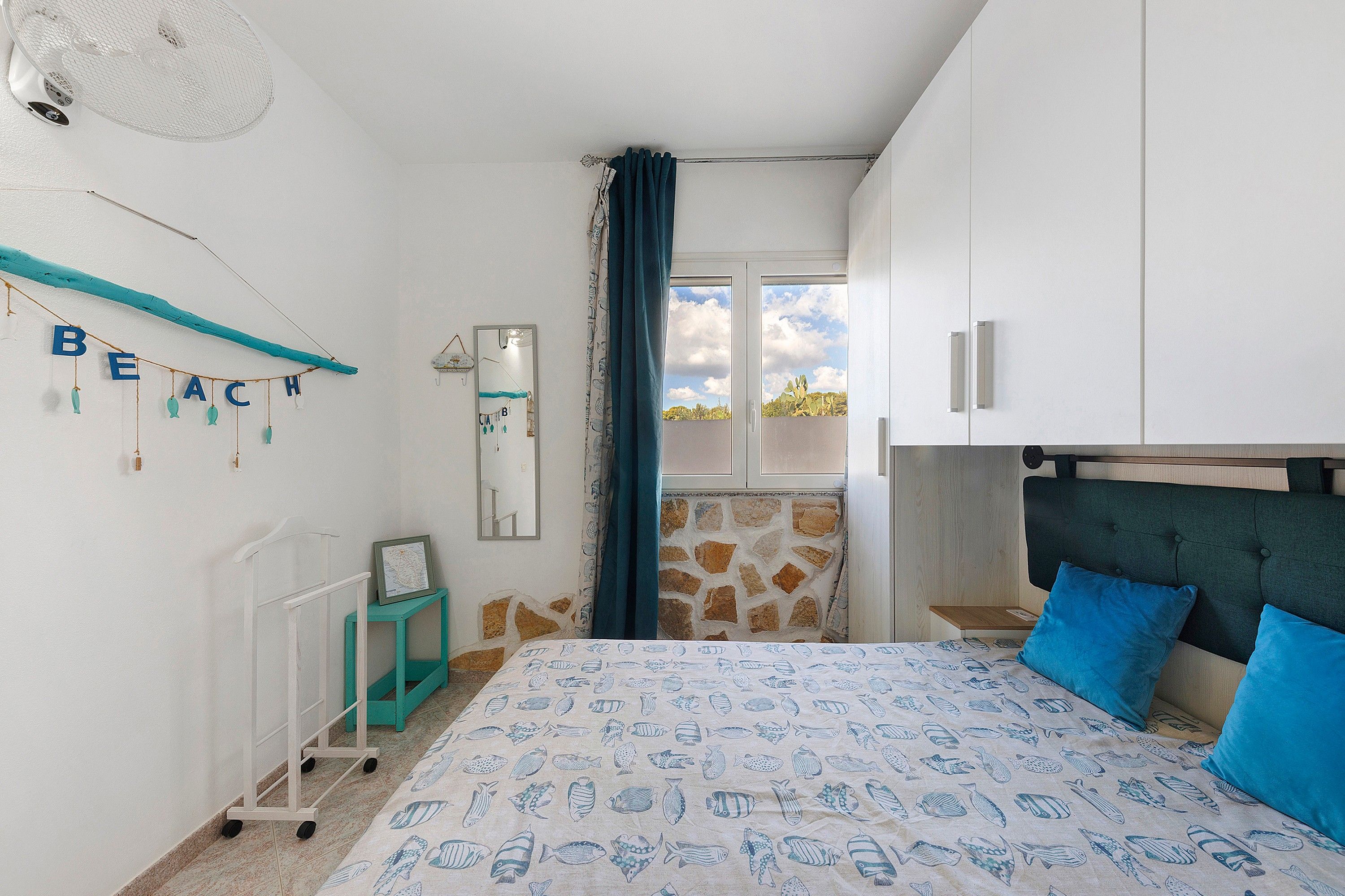 A bedroom with a bed, a dresser, a mirror, a map on a small table, and a wall with a 'BEACH' sign.