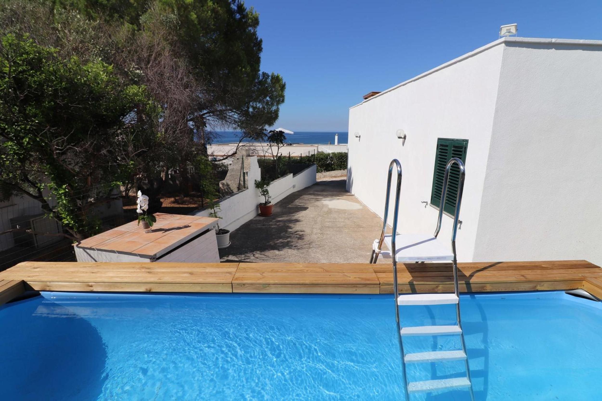A swimming pool with a wooden deck and a metal ladder, surrounded by a white building with a view of the sea.