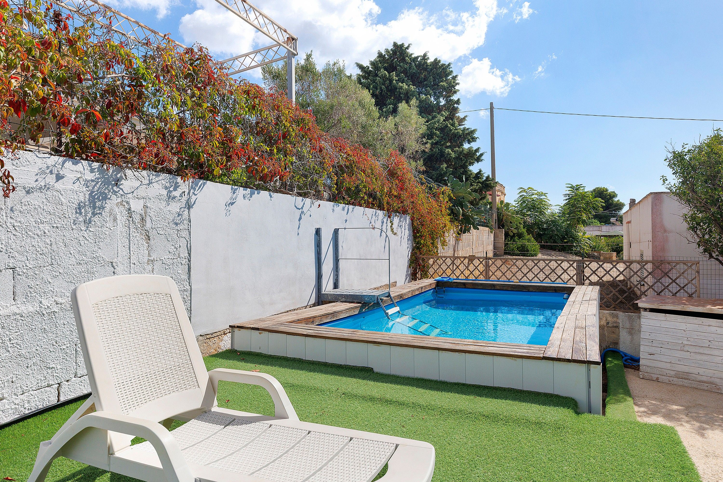 Ein Schwimmbecken mit Holzterrasse, einem weißen Stuhl und einer weißen Wand mit Kletterpflanzen.