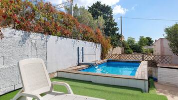 Ein Schwimmbecken mit Holzterrasse, einem weißen Stuhl und einer weißen Wand mit Kletterpflanzen.